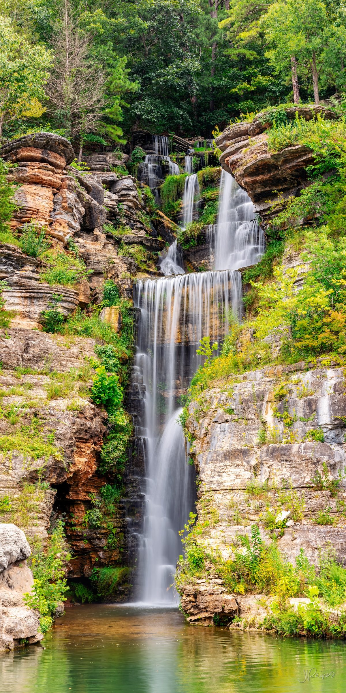 Multi-tiered waterfall cascading over layered limestone ledges into a green pool, framed by summer forest, Dogwood Canyon, Lampe, Missouri