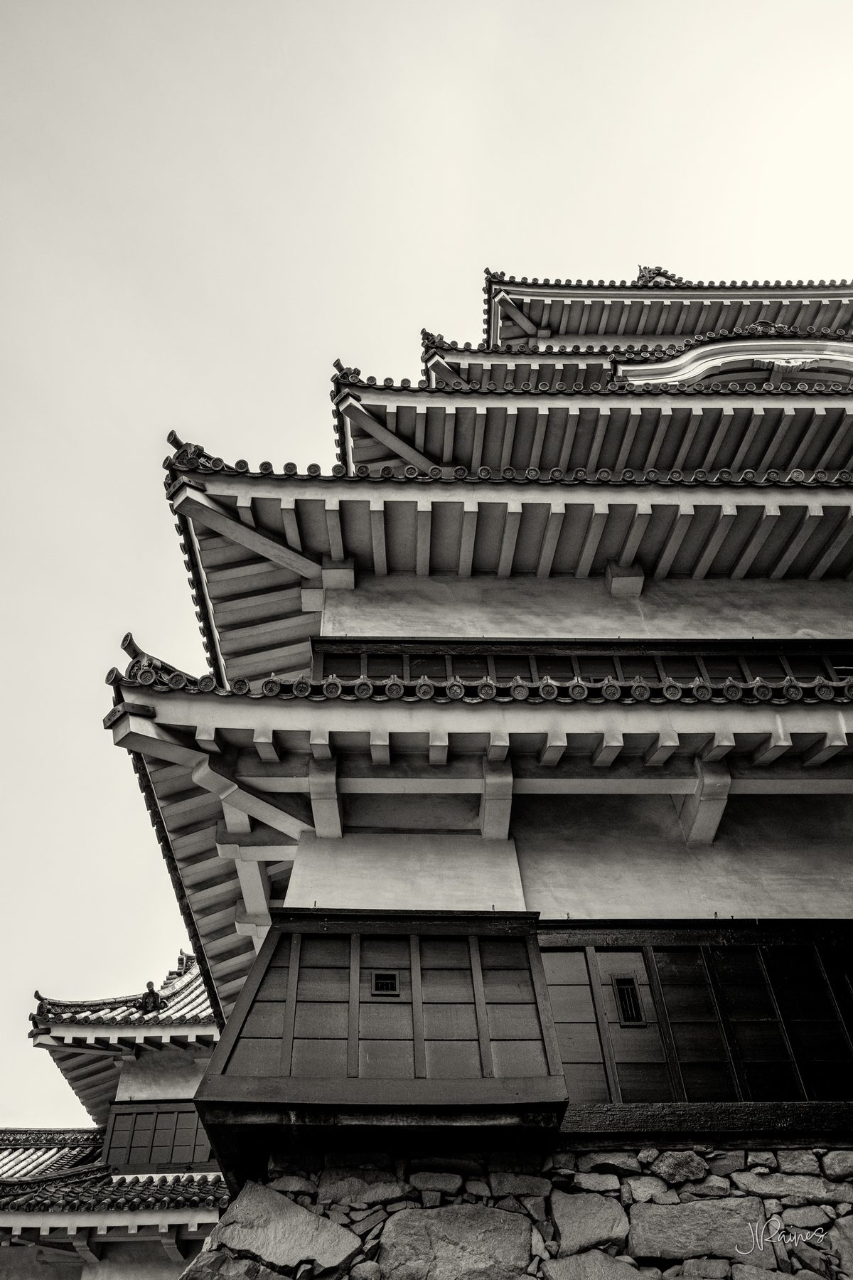 Steep low-angle view of Matsumoto Castle’s stacked black tiered roofs against open sky, in near-monochrome