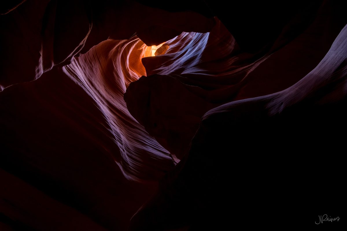 A heart-shaped slot canyon opening with warm glowing light at the far end and cool blue-lavender walls in shadow, Page, Arizona